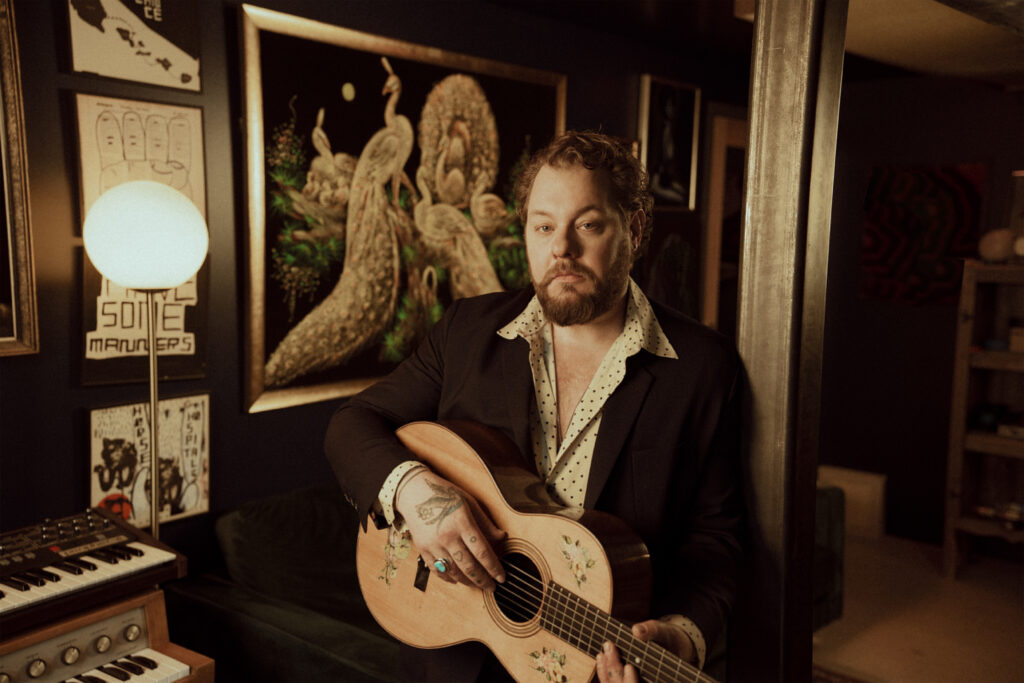 Musician Nathaniel Rateliff of Nathaniel Rateliff and the Nightsweats sitting in a moody room lit with orange and brown tones, holding a guitar.