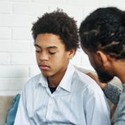 black man with short dreadlocks talking to a younger black male teen, both are sitting on a couch